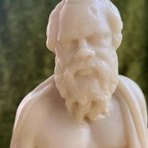 Könnte beinhalten: Eine cremefarbene Büste eines bärtigen Mannes mit Geheimratsecken, die detaillierte Gesichtszüge wie eine markante Nase, tief liegende Augen und einen strukturierten Bart aufweist und in ein einfaches Gewand über den Schultern gehüllt ist.