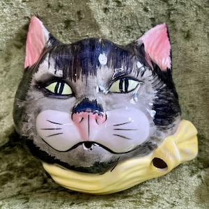 Puede incluir: Una cabeza de gato de cerámica con pelaje gris y negro, orejas rosas y ojos verdes. El gato tiene una nariz rosa y un lazo amarillo. Es un objeto decorativo.
