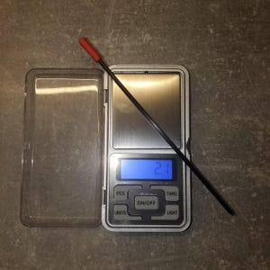 May include: A digital pocket scale with a silver metallic weighing platform and a blue backlit LCD screen displaying the number 2.1. A thin black rod with a red rubber tip is placed diagonally across the scale, which has a clear plastic cover.