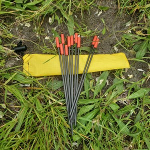 May include: A set of eight thin, dark gray metal tent stakes with bright orange plastic caps at the top ends. The stakes are arranged in a fan shape and are accompanied by a long, rectangular yellow storage pouch made of fabric.