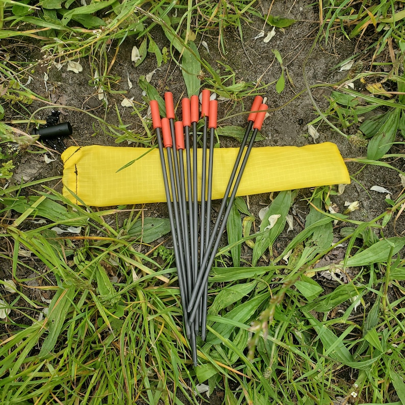 May include: A set of eight thin, dark gray metal tent stakes with bright orange plastic caps at the top ends. The stakes are arranged in a fan shape and are accompanied by a long, rectangular yellow storage pouch made of fabric.