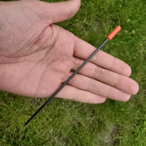 May include: A thin, straight, black metal rod featuring a small, cylindrical, bright orange cap at one end. The rod is slender and uniform in thickness, with a pointed tip at the opposite end from the orange cap, presenting a simple, functional design.