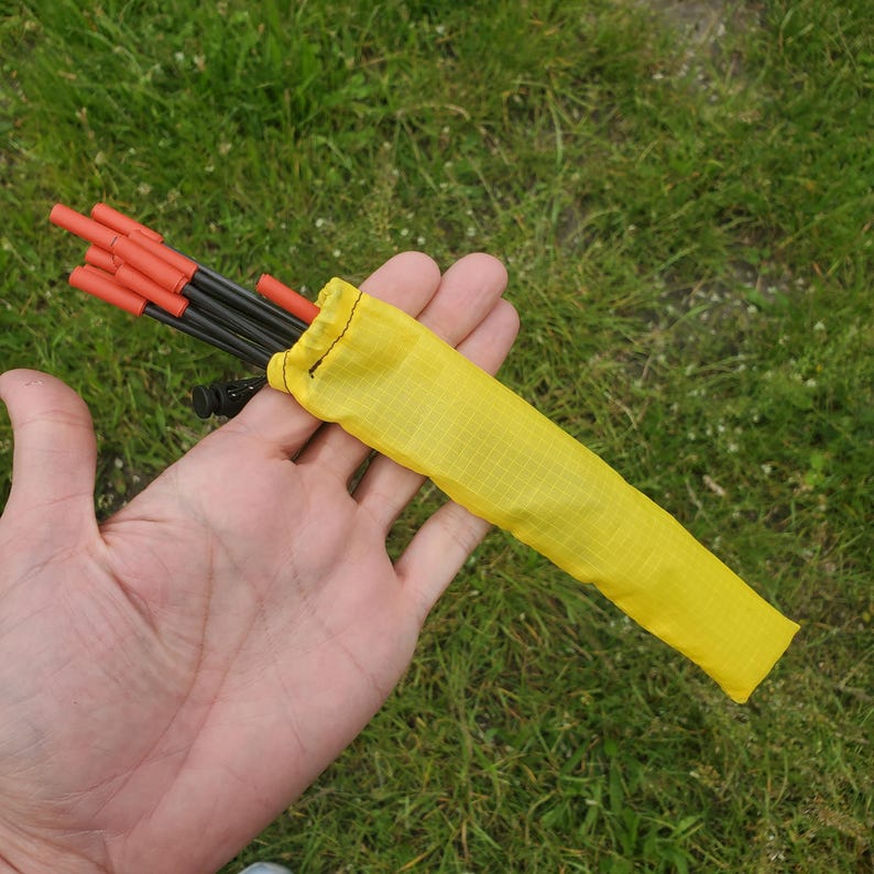 May include: A set of thin black metal tent stakes with bright orange tips, partially inserted into a yellow ripstop nylon storage pouch with a drawstring closure. The stakes are bundled together and held in a hand against a grassy background.