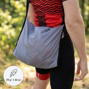 May include: A lightweight grey crossbody bag with a black strap, worn over a red and black zigzag patterned shirt and black shorts. A circular white icon displays a feather graphic with the text 29 g | 1.02 oz, indicating the weight of the bag.