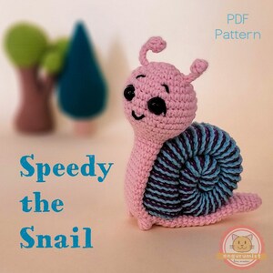 May include: A pink and blue crocheted snail amigurumi toy named "Speedy the Snail". The snail has a smiling face with black eyes and a blue and purple striped shell.