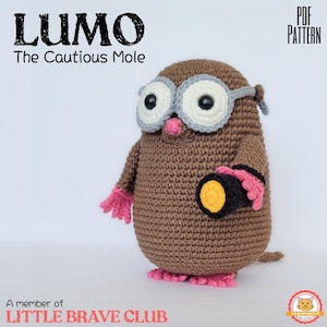May include: A brown crocheted mole toy named Lumo the Cautious Mole, featuring large white and grey goggles, a small pink nose, pink hands and feet, and holding a black flashlight with a yellow light. Text reads PDF PATTERN and LITTLE BRAVE CLUB.