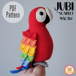 Könnte beinhalten: Gehäkelter roter Ara-Papagei mit roten, gelben, orangefarbenen, grünen und blauen Federn. Der Vogel hat ein weißes und schwarzes Auge, einen beigen Schnabel und eine graue Sitzstange. Der Text "PDF Pattern" und "Jubi the Scarlet Macaw" sind sichtbar.