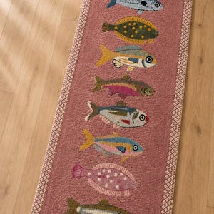 Könnte beinhalten: Ein rechteckiger Läufer in Altrosa mit einer vertikalen Reihe von neun gestickten Fischen in verschiedenen Farben wie Gelb, Blau, Grün und Rot. Der Teppich hat eine strukturierte Oberfläche, eine cremefarbene geometrische Bordüre und Fransen.