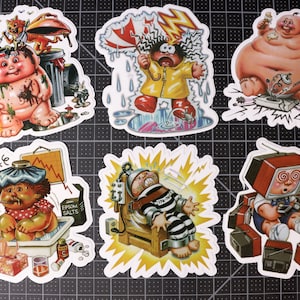 May include: Six colorful stickers featuring cartoon characters with various expressions and situations. The characters are all babies with exaggerated features. The stickers are printed on white paper with a white border.