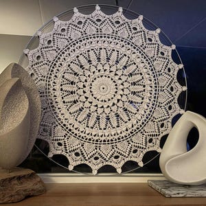 Könnte beinhalten: Ein großer, kreisförmiger, weiß gehäkelter Deckchen mit einem detaillierten Spitzenmuster wird gezeigt. Zwei abstrakte Skulpturen befinden sich davor. Eine ist eine hellgraue, gebogene Form, die andere eine weiße, tropfenförmige Skulptur.