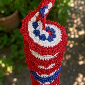 May include: A spiral-shaped crochet hanging decoration featuring a repeating pattern of red, white, and blue yarn. The structure consists of interconnected circular segments with a scalloped red border, suspended by a single red crochet loop at the top.