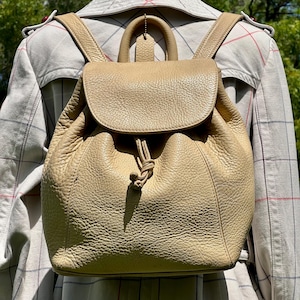 May include: A tan leather backpack with a flap closure and drawstring. The backpack has adjustable shoulder straps and a top handle. The leather has a textured appearance. The background includes a plaid trench coat.