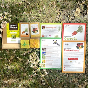 May include: A Mud & Bloom gardening box containing organic carrot and nasturtium seeds, two yellow paper bundles, wooden craft sticks, and instructional cards for growing vegetables and identifying insects, all arranged on a grassy surface.