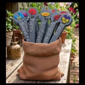 May include: A collection of grey garden plant markers with colourful floral icons and white text labels including Sweet Pea, Poppy, Sunflower, Snapdragon, and Pansy, all held within a brown, textured, sack-shaped ceramic container.