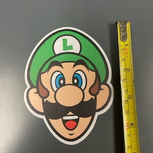 Può includere: Un adesivo di un personaggio dei cartoni animati verde e marrone con un grande viso amichevole. Il personaggio indossa un cappello verde con una L sopra, un baffo marrone e un grande sorriso.