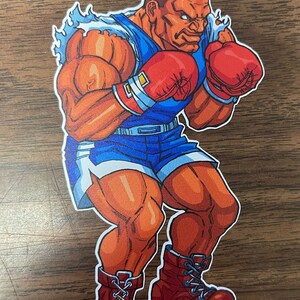 Puede incluir: Una ilustración de dibujos animados de un boxeador musculoso con guantes de boxeo rojos, pantalones cortos azules y botas rojas. El boxeador tiene una expresión decidida en su rostro.