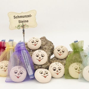 Könnte beinhalten: Eine Sammlung von lächelnden Gesichtssteinen, die jeweils mit einem fröhlichen Ausdruck handbemalt wurden. Die Steine werden auf einer kleinen Felsformation und in farbigen Organzabeuteln präsentiert. Ein Schild trägt die Aufschrift "Schmunzel-Steine".