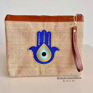 Può includere: Pochette beige con finiture e cinturino da polso in pelle marrone. La pochette presenta una mano di Hamsa ricamata in blu con un design a occhio diabolico. La cerniera e l'hardware sono dorati. Il testo "@Makrameeinlove By HILAL" è visibile.