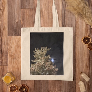 Op de afbeelding: Een witte canvas tote bag met een zwart-witte afbeelding van een boom die 's nachts met lichtjes is verlicht. De boom staat voor een donkere hemel met een zichtbare maan.