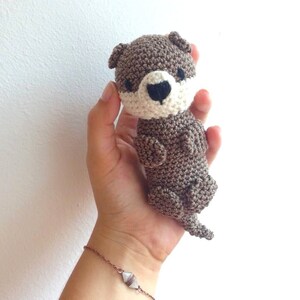 Könnte beinhalten: Ein gehäkeltes Plüschtier einer Otter mit braunem Fell, weißen Gesichtsdetails und schwarzen Augen. Das Spielzeug wird in der Hand einer Person gehalten.