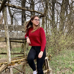 Peut inclure: Une femme portant un pull en tricot rouge, un jean noir et des baskets blanches est assise sur une structure en bois dans un environnement forestier.