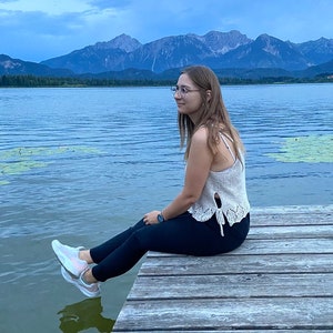 Puede incluir: Una persona con un top de crochet blanco y pantalones negros se sienta en un muelle de madera con vistas a un lago con monta&ntilde;as en el fondo.