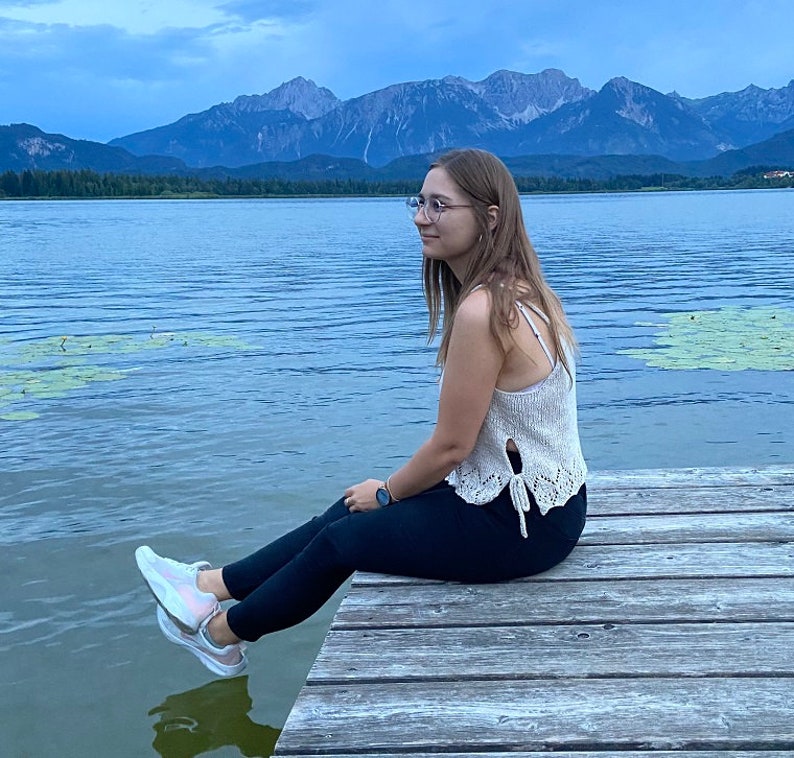 Puede incluir: Una persona con un top de crochet blanco y pantalones negros se sienta en un muelle de madera con vistas a un lago con monta&ntilde;as en el fondo.