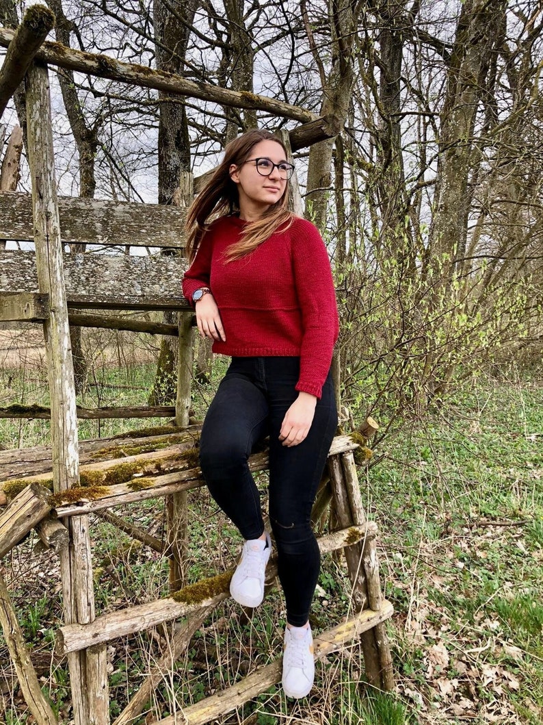 May include: A woman wearing a red knitted sweater, black jeans, and white sneakers sits on a wooden structure in a forest setting.
