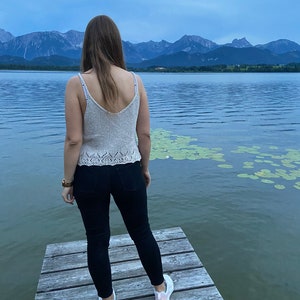 Puede incluir: Una persona con un top de punto beige claro y pantalones vaqueros negros se encuentra en un muelle de madera con vistas a un lago con monta&ntilde;as en el fondo.