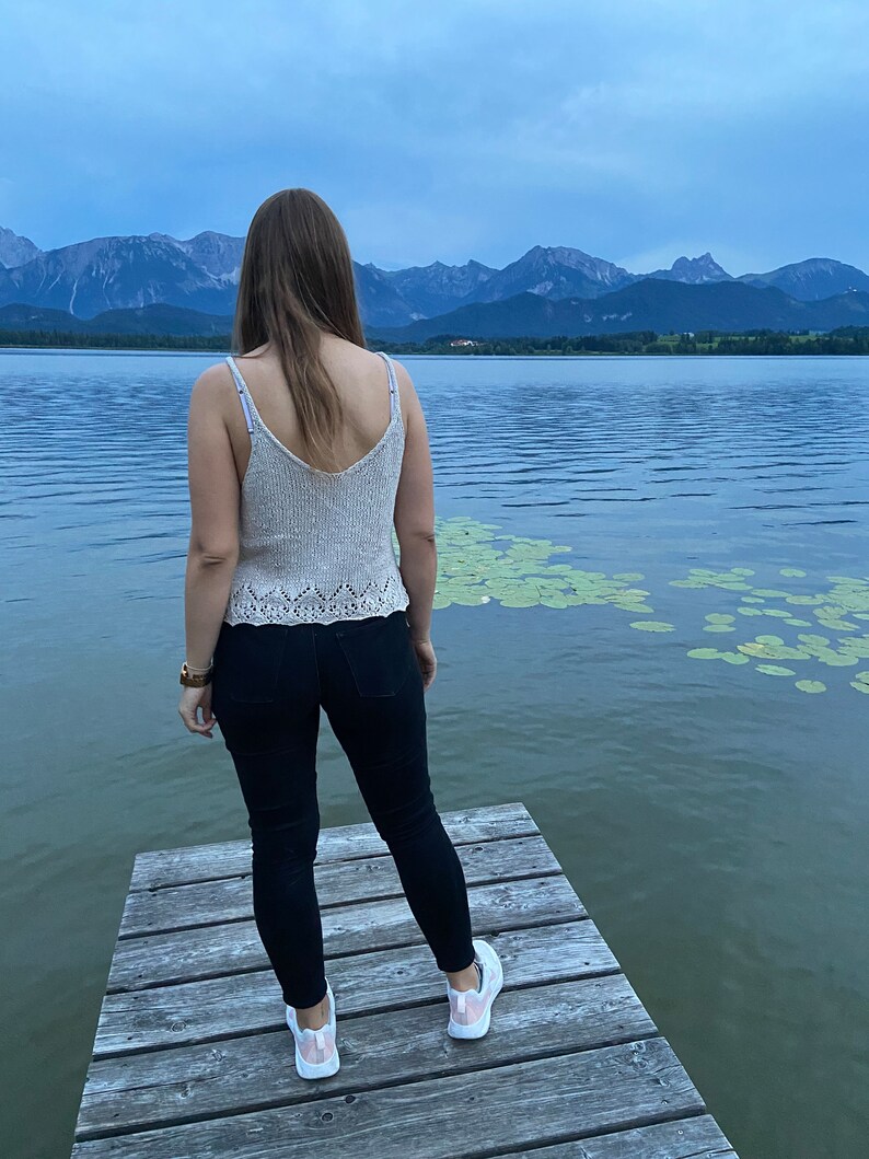 Puede incluir: Una persona con un top de punto beige claro y pantalones vaqueros negros se encuentra en un muelle de madera con vistas a un lago con monta&ntilde;as en el fondo.