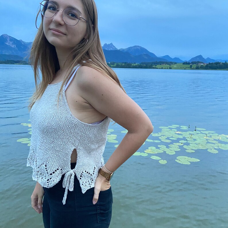 Puede incluir: Una mujer lleva un top de crochet beige claro con un detalle de nudo en la parte inferior. Est&aacute; de pie junto a un lago con monta&ntilde;as en el fondo.