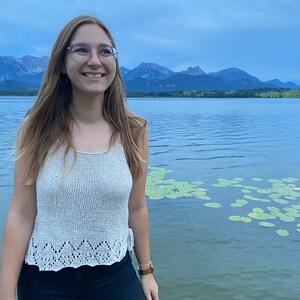 Puede incluir: Una mujer con un top blanco de crochet con un delicado borde de encaje, pantalones negros y un reloj marr&oacute;n. Est&aacute; de pie frente a un lago con monta&ntilde;as en el fondo.