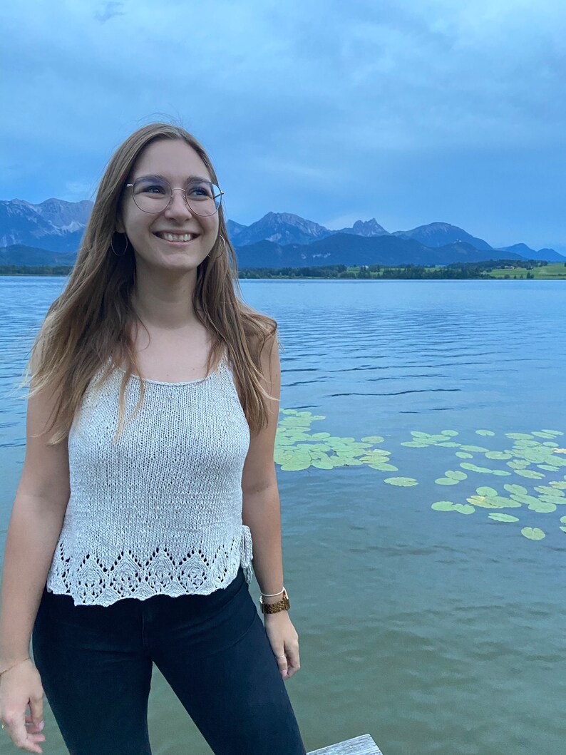 Puede incluir: Una mujer con un top blanco de crochet con un delicado borde de encaje, pantalones negros y un reloj marr&oacute;n. Est&aacute; de pie frente a un lago con monta&ntilde;as en el fondo.