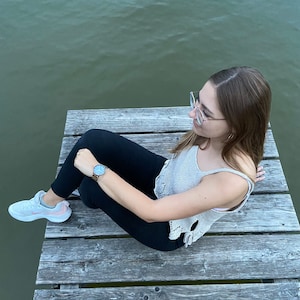 Puede incluir: Una persona con un top de crochet blanco, pantalones negros y zapatillas blancas se sienta en un muelle de madera sobre una masa de agua. La persona lleva un reloj en la mu&ntilde;eca izquierda y gafas en la cara.