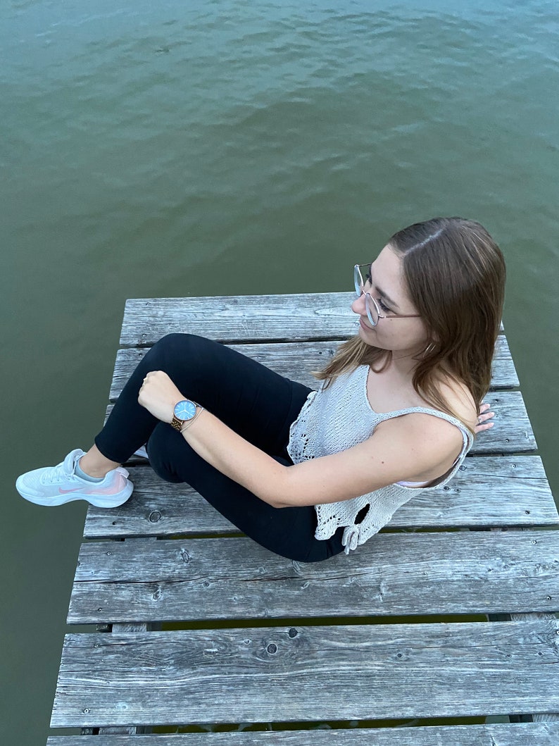 Puede incluir: Una persona con un top de crochet blanco, pantalones negros y zapatillas blancas se sienta en un muelle de madera sobre una masa de agua. La persona lleva un reloj en la mu&ntilde;eca izquierda y gafas en la cara.