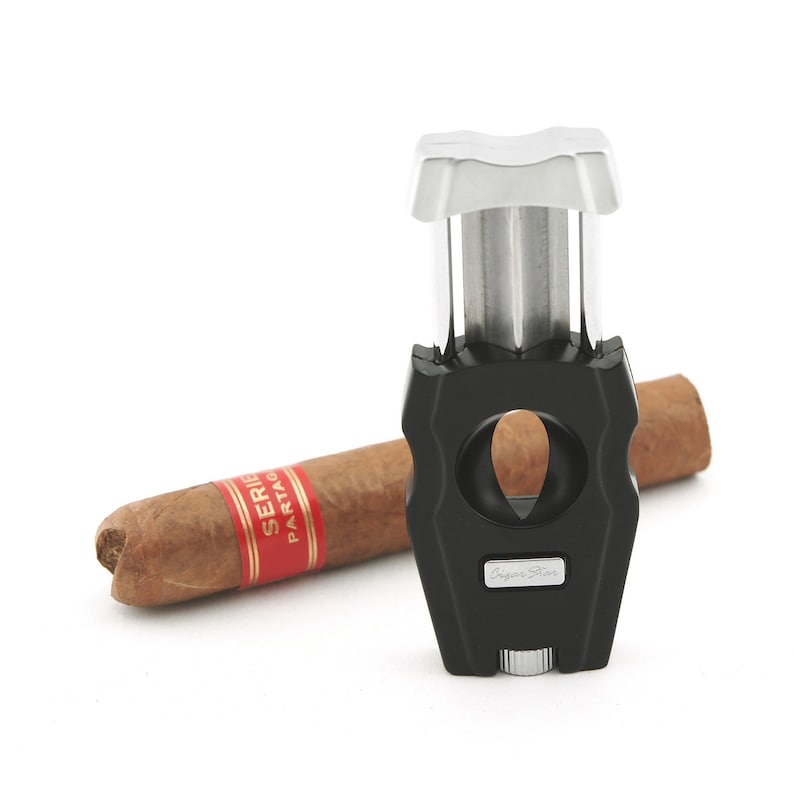 May include: A black and silver cigar cutter with a guillotine blade. The cutter is next to a brown cigar with a red and gold band that says "Serie  Partagas".