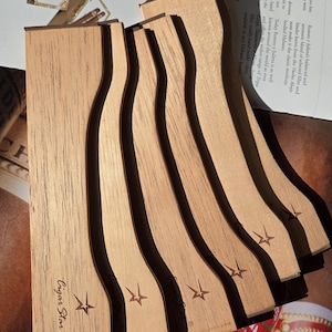 Puede incluir: Cinco reposacigarros de madera con un diseño de estrella y el texto "Cigar Star" en la parte inferior. Los reposacigarros están apilados uno encima del otro.