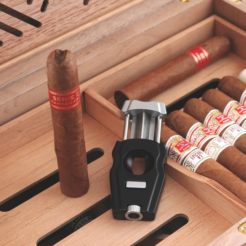 May include: A wooden humidor with a black cigar cutter and several cigars. The cigars are labelled "Serie D" and "Ep&iacute;cure No. 2".