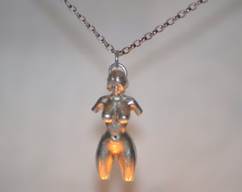 Könnte beinhalten: Halskette mit silberfarbenem Anhänger, der eine stilisierte menschliche Figur darstellt. Der Anhänger ist eine dreidimensionale Skulptur eines Torso, der an einer feinen Kette hängt. Die Figur hat eine glatte, polierte Oberfläche vor neutralem Hintergrund.
