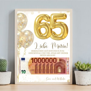 Op de afbeelding: Een ingelijste verjaardagsafdruk met gouden folieballonnen en het getal 65 in goud. De afdruk bevat de tekst "Liebe Maria!" en een afbeelding van eurobankbiljetten ter waarde van 1.000.000. De afdruk is versierd met gouden confetti.