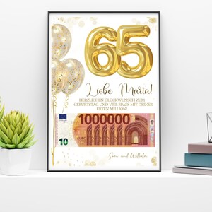 Op de afbeelding: Een ingelijste print met een witte achtergrond toont gouden folieballonnen en het nummer "65" in goud. De tekst "Liebe Maria!" staat boven een afbeelding van eurobiljetten. De print zit in een zwarte lijst.