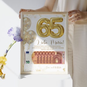 Op de afbeelding: Een ingelijste print met gouden folieballonnen die het getal "65" vormen en de tekst "Liebe Maria!". De print bevat een afbeelding van een biljet van 10 euro en de tekst "1000000". De print zit in een licht houten frame.