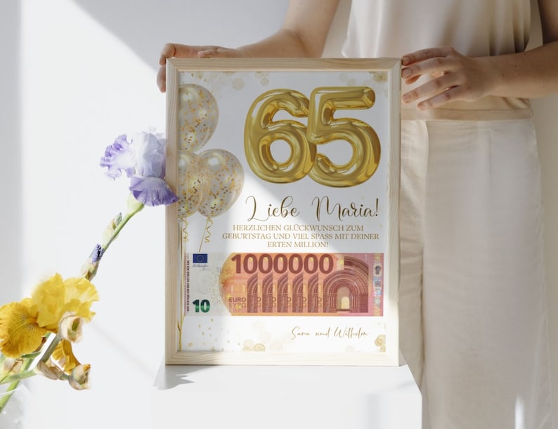 Op de afbeelding: Een ingelijste print met gouden folieballonnen die het getal "65" vormen en de tekst "Liebe Maria!". De print bevat een afbeelding van een biljet van 10 euro en de tekst "1000000". De print zit in een licht houten frame.