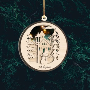 May include: A wooden Christmas ornament featuring a laser-cut design of a snowy village scene with a church, street lamp, and snowman. The ornament is round and has a brown border. The text "Let it snow" is written on the bottom of the ornament.