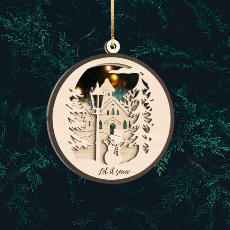 May include: A wooden Christmas ornament featuring a laser-cut design of a snowy village scene with a church, street lamp, and snowman. The ornament is round and has a brown border. The text "Let it snow" is written on the bottom of the ornament.