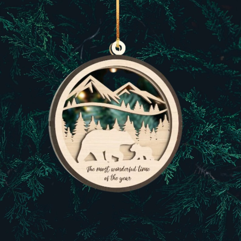 May include: Wooden Christmas ornament featuring a silhouette of a mother bear and cub in a forest setting with mountains in the background. The ornament has the text "The most wonderful time of the year" inscribed on the bottom.