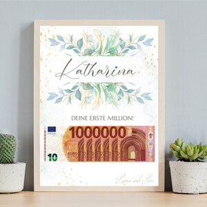 May include: A framed print with a watercolor illustration of green leaves and gold accents. The text "Katharina" is written in a decorative font. The text "Deine erste Million!" is written below the illustration. A mock-up of a 100,000 euro bill is shown below the text. The text "Laura und Tom" is written at the bottom of the print.