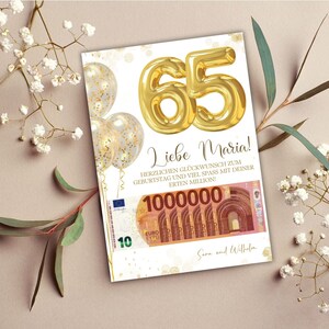Op de afbeelding: Een verjaardagskaart met gouden folieballonnen die het getal 65 vormen. De kaart bevat de tekst "Liebe Maria!" en een afbeelding van een biljet van 100 euro. Gouden confetti-ballonnen en bloemaccenten maken het ontwerp compleet.