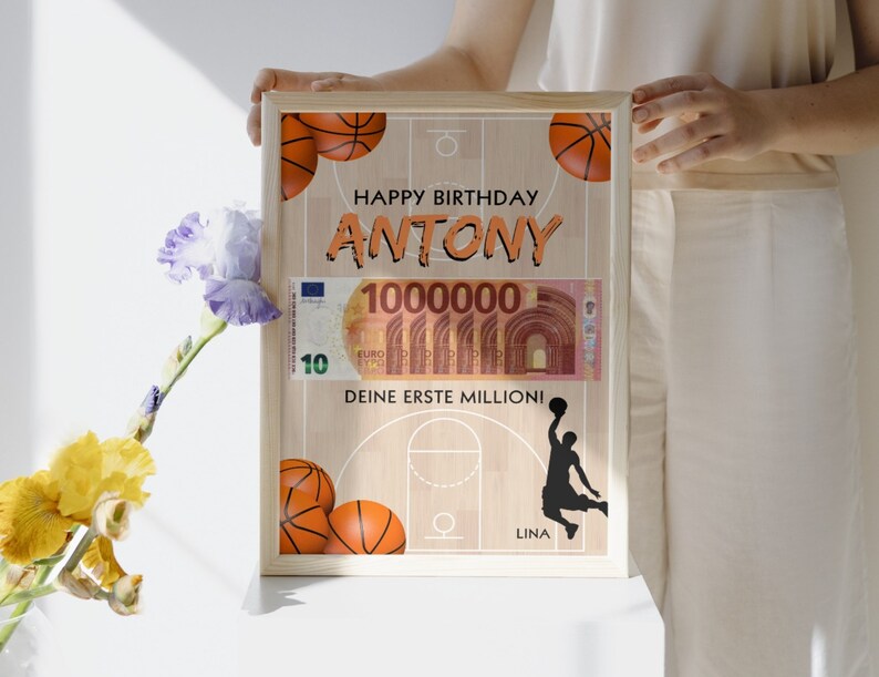 Puede incluir: Una tarjeta de cumplea&ntilde;os enmarcada con tem&aacute;tica de baloncesto. La tarjeta presenta una cancha de baloncesto con un aro de baloncesto, balones de baloncesto y una silueta de un jugador de baloncesto. La tarjeta tambi&eacute;n incluye un billete de dinero falso con el texto "100000" y las palabras "Happy Birthday Antony, Deine erste Million!" y "Lina".