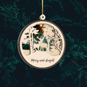 May include: A wooden Christmas ornament with a laser-cut design of two deer in a forest scene. The ornament is brown with a light wood interior and has the text "Merry and Bright" written on the bottom.